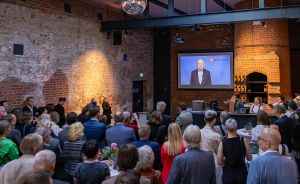 Publikum in Veranstaltungshalle mit Backsteinwänden schaut auf Leinwand mit sprechendem Mann im Anzug.