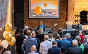 Mann spricht vor Publikum in Veranstaltungsraum mit Backsteinwand, Leinwand mit orangenen Kugeln und gold-weißen Luftballons.