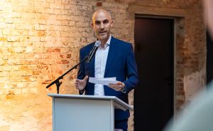 Mann im blauen Anzug spricht an einem Rednerpult mit Mikrofon, hält Papier in der Hand, Backsteinwand im Hintergrund.