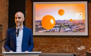 Mann im blauen Sakko spricht an einem Podium mit Mikrofon, hinter ihm eine Leinwand mit orangefarbenen Kugeln.