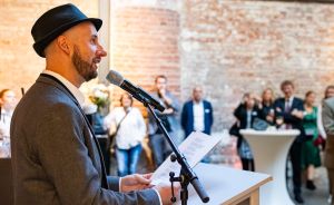Mann mit schwarzem Hut und grauem Jacket spricht an Mikrofon am Podium, Publikum im Hintergrund.