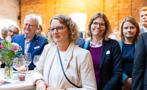 Mehrere Personen in Business-Kleidung stehen bei einer Veranstaltung in einem Raum mit Backsteinwand und Stehtischen.