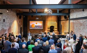 Publikum in Industriehalle mit Backsteinwänden, Person am Podium, Bildschirm mit orangem Ballon, Diskokugel an der Decke.