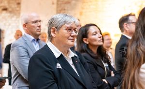 Mehrere Personen in formeller Kleidung stehen in einem Raum mit Steinwänden und hören aufmerksam zu.