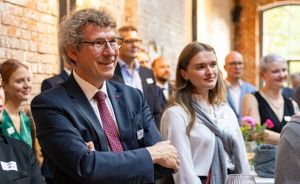 Mann mit Brille und Krawatte lächelt mit verschränkten Armen; dahinter stehen mehrere Personen in einem Raum.