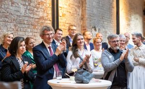 Gruppe von Menschen applaudiert in einem Veranstaltungsraum mit Backsteinwand, Stehtischen und festlicher Atmosphäre.