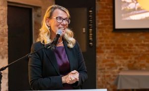 Frau mit blonden Haaren und Brille spricht lächelnd an einem Mikrofon, gekleidet in schwarzem Blazer und lila Oberteil.