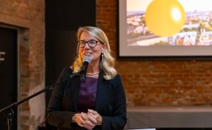 Frau mit Brille und blonden Haaren spricht an einem Mikrofon vor einer Präsentationsleinwand in einem Backsteinraum.