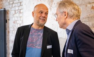 Zwei ältere Männer mit Namensschildern unterhalten sich vor einer Backsteinwand.