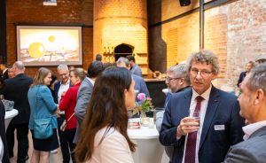 Personen in Businesskleidung beim Netzwerken in einem Veranstaltungsraum mit Backsteinwänden und Präsentationsleinwand.