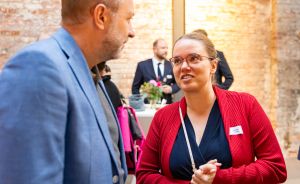 Zwei Personen im Gespräch bei einer Veranstaltung; Frau in roter Jacke mit Namensschild, Mann im blauen Blazer.