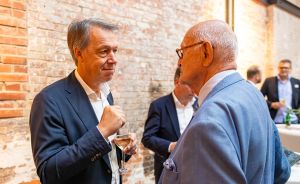 Zwei ältere Männer in Anzügen unterhalten sich bei einer Veranstaltung; einer hält ein Weinglas. Backsteinwand im Hintergrund.