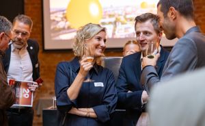 Drei Personen mit Weingläsern im Gespräch bei einer Veranstaltung, im Hintergrund eine Präsentation und weitere Gäste.