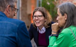 Drei Personen sitzen zusammen im Gespräch. Eine Frau mit Brille lacht. Zwei weitere Personen sind von hinten zu sehen.