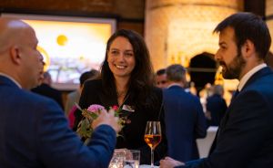 Drei Personen bei einer Veranstaltung, eine Frau mit Weinglas lächelt, zwei Männer in Anzügen, Blumen auf dem Tisch.
