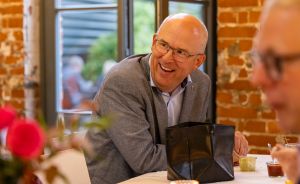 Mann mit Brille und grauem Sakko lacht an einem Tisch in einem Restaurant mit Backsteinwänden.