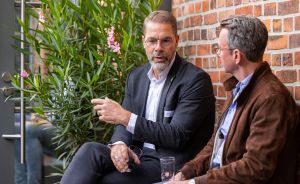 Zwei Männer mit Brille sitzen vor Backsteinwand und Pflanze im Gespräch; einer gestikuliert, beide tragen Jackett.