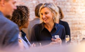 Drei Personen unterhalten sich bei einem Event. Eine blonde Frau hält ein Glas und lächelt. Backsteinwand im Hintergrund.