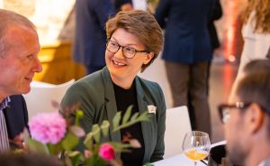 Frau mit kurzen Haaren und Brille im grünen Blazer sitzt lächelnd an einem Tisch mit Blumen und Weinglas.