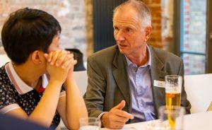 Zwei Personen sitzen an einem Tisch im Gespräch. Ein älterer Mann mit Namensschild gestikuliert, vor ihm steht ein Bier.