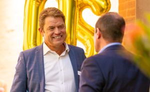 Zwei Männer in blauen Anzügen im Gespräch, im Hintergrund goldene Zahlenballons an einer Backsteinwand.