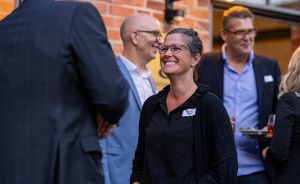 Frau mit Brille und schwarzem Hemd lächelt bei einer Networking-Veranstaltung, umgeben von Personen in Businesskleidung.