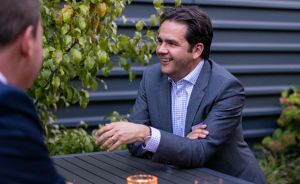 Zwei Männer sitzen an einem Tisch im Freien. Ein Mann im grauen Anzug lächelt und gestikuliert beim Gespräch.