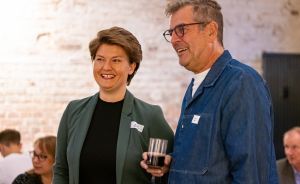 Zwei Personen mit Namensschildern lachen in einem Raum mit Backsteinwand. Mann hält ein Glas.