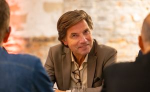 Mann mittleren Alters im Sakko sitzt lächelnd an einem Tisch und spricht mit zwei Personen in einem Café.