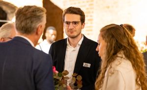 Mann mit Brille und Namensschild im Gespräch mit zwei Personen bei einer Veranstaltung, Backsteinwand im Hintergrund.