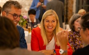 Frau in rotem Blazer lächelt bei einer Veranstaltung. Um sie herum sitzen mehrere Personen an einem Tisch.