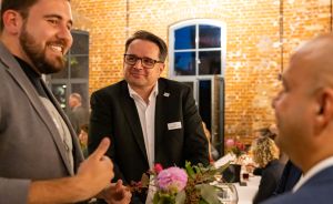 Drei Männer im Gespräch bei einer Abendveranstaltung, Stehtisch mit Blumen, Backsteinwand im Hintergrund.