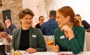 Zwei Frauen mit Namensschildern sitzen an einem Tisch, lachen, eine hält ein Weinglas. Im Hintergrund weitere Personen.