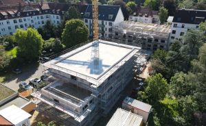Luftaufnahme einer Baustelle mit Gerüst und Kran, umgeben von Wohngebäuden und Bäumen.