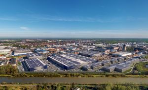 Luftaufnahme eines großen Industriegeländes mit Lagerhallen und Solardächern an einem Fluss, dahinter eine Stadt.