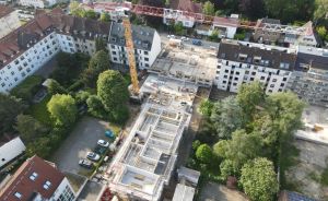 Luftaufnahme einer Baustelle mit Kran zwischen mehrstöckigen Wohngebäuden, umgeben von Bäumen in städtischer Umgebung.