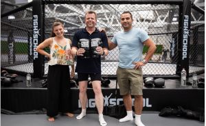 Drei Personen stehen lächelnd vor einem MMA-Käfig mit "Fight School"-Beschriftung; der mittlere hält Boxhandschuhe.