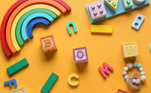 Bunte Holzspielzeuge auf orangem Hintergrund: Regenbogen, Buchstabenblöcke (B, O, P, M, C, S, J), Bausteine und Beißring.
