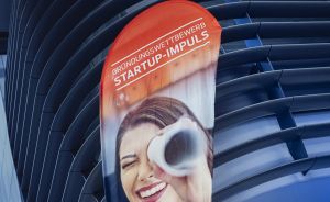 Werbebanner mit Aufschrift „Gründungswettbewerb Startup-Impuls" und lachender Frau mit Kaffeetasse, vor dunkler Gebäudefassade.