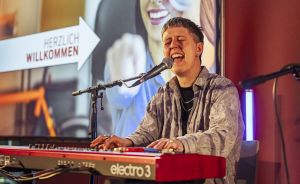 Person spielt rotes Nord Electro 3 Keyboard und singt ins Mikrofon. Im Hintergrund Schild „Herzlich Willkommen".