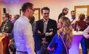 Drei Personen im Gespräch bei einer Abendveranstaltung, im Hintergrund weitere Gäste an Stehtischen mit Getränken.