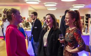 Drei Frauen unterhalten sich lachend auf einer Veranstaltung; eine hält ein Weinglas. Im Hintergrund weitere Gäste.