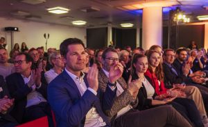 Publikum klatscht in einem beleuchteten Veranstaltungssaal; im Vordergrund ein Mann im blauen Sakko.