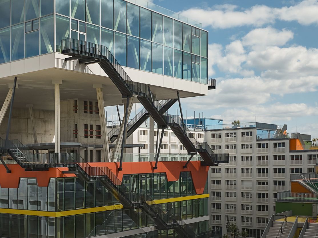Modernes Gebäude mit Glasaufbau auf Stahlstützen, orangefarbenen Elementen und Außentreppen, dahinter mehrstöckige Bürogebäude.