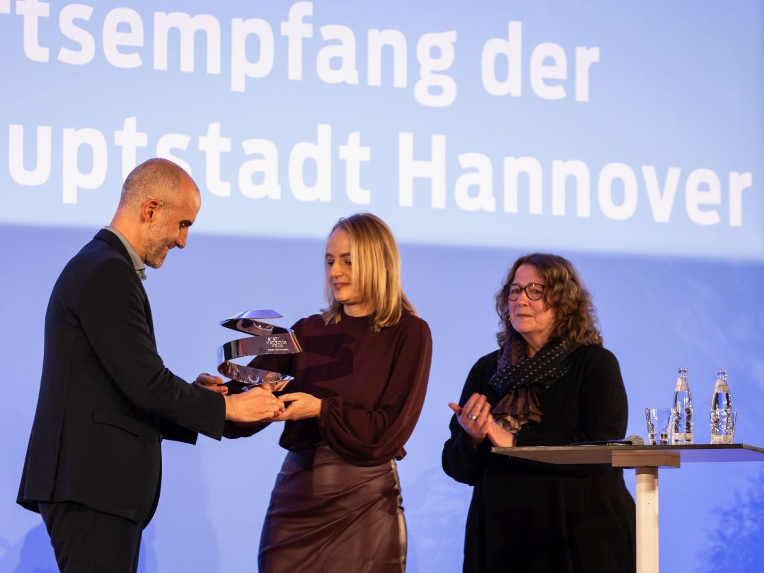 Drei Personen bei einer Preisverleihung in Hannover. Ein Mann überreicht einer Frau eine Auszeichnung, eine zweite Frau applaudiert.