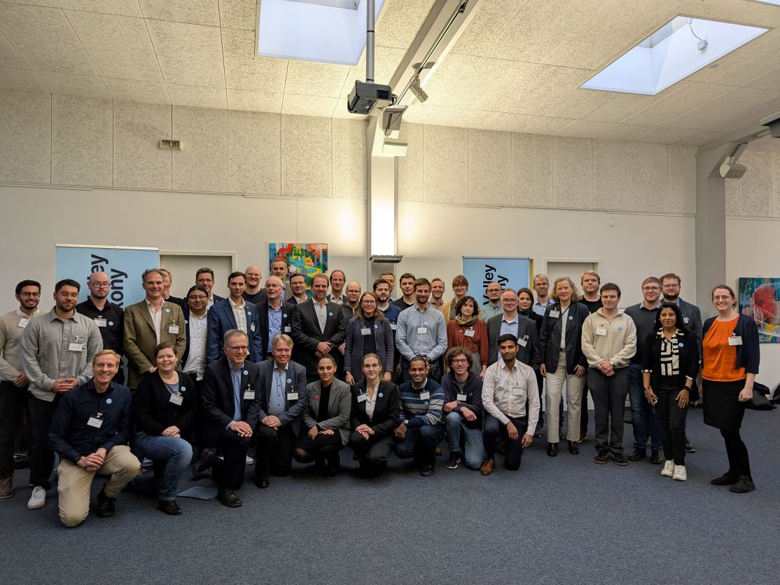 Große Gruppe von etwa 40 Personen posiert für ein Gruppenfoto in einem Konferenzraum mit blauen Aufstellern.