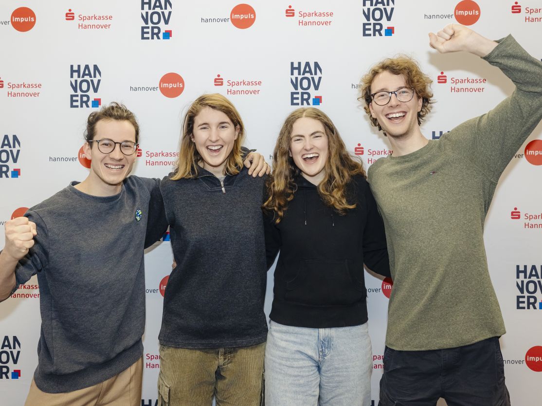 Vier junge Menschen stehen lachend vor einer Sponsorenwand mit Logos von Sparkasse Hannover und Hannover Impuls.