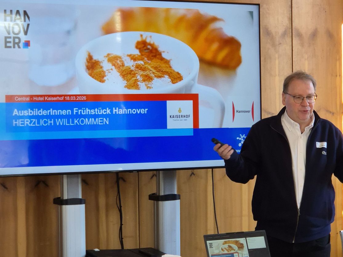 Mann steht vor Präsentationsfolie „AusbilderInnen Frühstück Hannover – Herzlich Willkommen" im Hotel Kaiserhof.