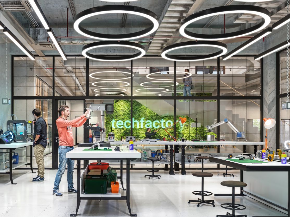 Modernes Labor "techfactory" mit drei Personen an Arbeitstischen, Ringlampen an der Decke und grüner Pflanzenwand.