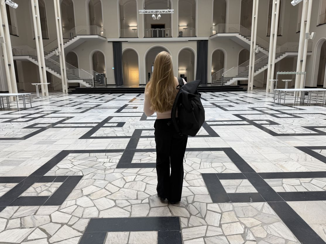 Frau mit langen blonden Haaren und schwarzem Rucksack steht in einer großen, hellen Halle mit gemustertem Marmorboden.
